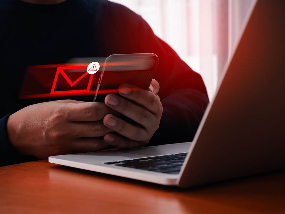 Person holding a phone beside a laptop with a warning email graphic