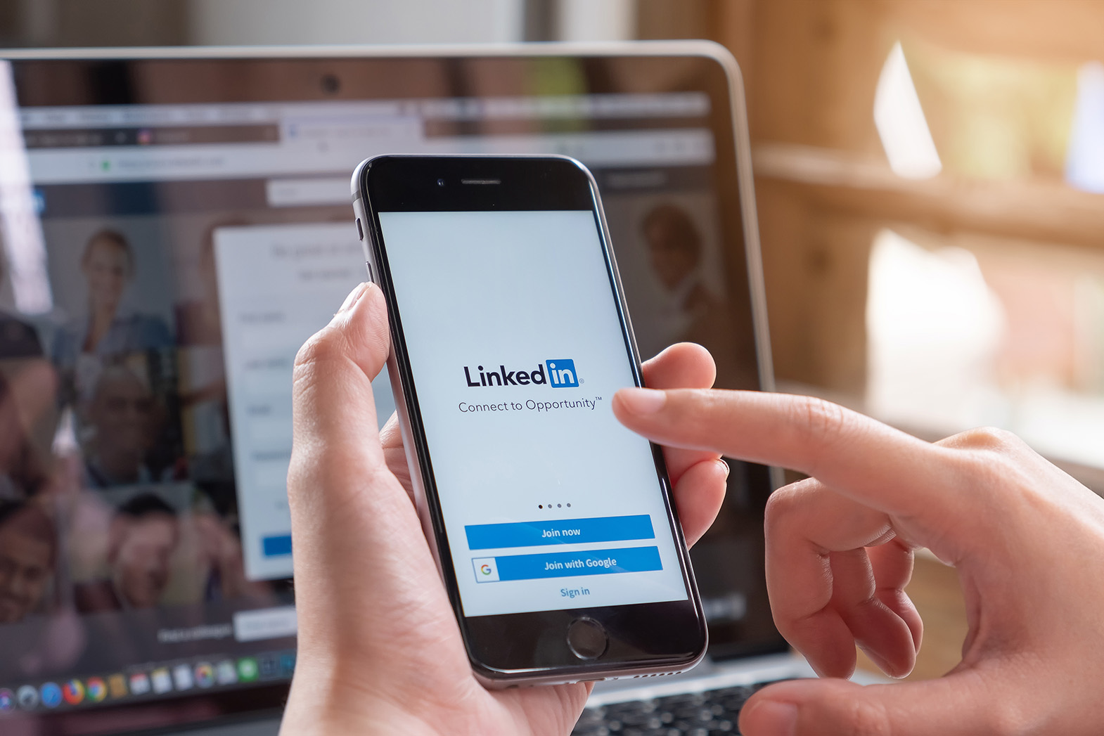 Hands holding a phone displaying LinkedIn in front of a laptop