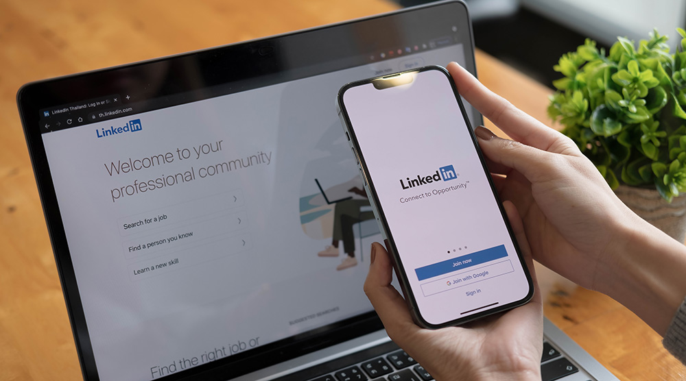 Person holding a phone with LinkedIn open beside a laptop showing LinkedIn