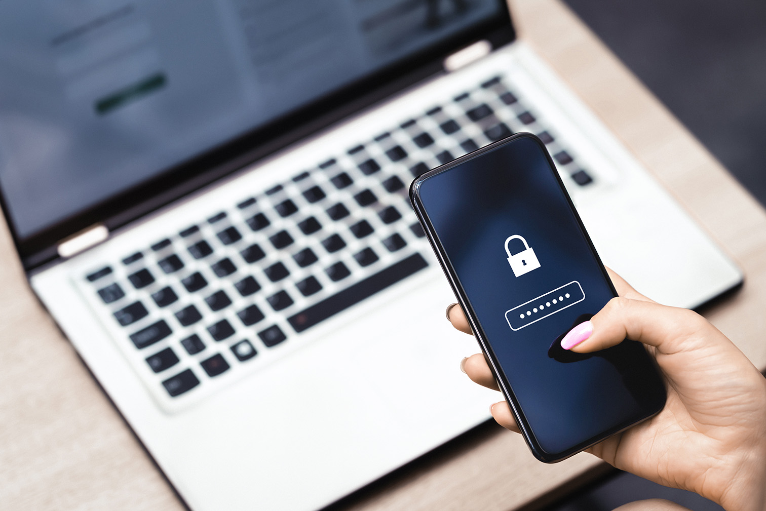 Hand holding a phone with a padlock and masked password above a laptop